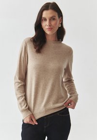 Beige gebreid sweater met een ronde halslijn, lange mouwen en ribbelboorden aan de mouwen en onderkant, gecombineerd met donkere jeans. Zachte textuur, minimalistisch ontwerp.