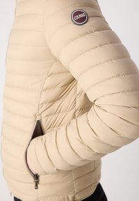 Beige puffjacka med quiltat mönster, sidofickor med dragkedja och rund logotyppatch på ärmen. Slät, lättviktig material.