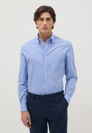 Jeune homme aux cheveux bruns portant une chemise bleue claire à boutons et un pantalon bleu foncé, debout devant un fond blanc uni.