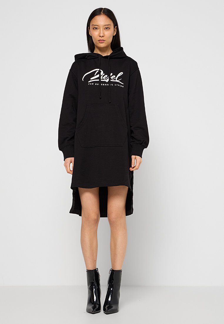 Zwarte hoodie jurk met lange mouwen, een voorzak en zijsplitten. Voorzien van witte grafische tekst op de borst. De stof lijkt zacht en gestructureerd.