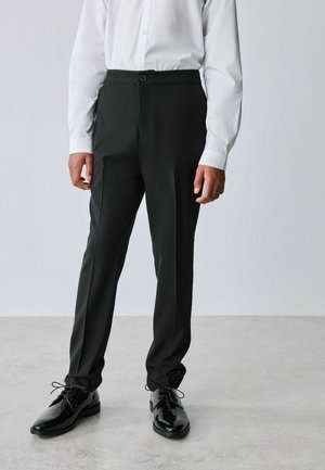 Mand iført sorte bukser, hvid skjorte med knapper og sorte polerede sko, stående på lys grå gulv mod en ensfarvet baggrund.