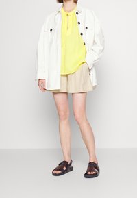 Bílá oversized košile, jasně žlutý top, béžové kraťasy a tmavě hnědé sandály s detaily na popruzích. Minimalistický design, neformální outfit.