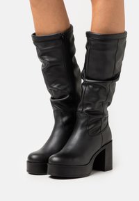 Stivali neri al ginocchio in pelle liscia, con zip laterale, design morbido, tacco alto spesso e suola piatta.