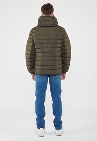 Giacca imbottita verde oliva con design a righe orizzontali, cappuccio e polsini elasticizzati. Indossata sopra jeans blu e sneakers bianche.
