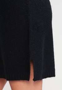 Svart stickad klänning med texturerad tyg, med en sidoklyvning och ribbad fåll. Materialet verkar mjukt och något fluffigt.