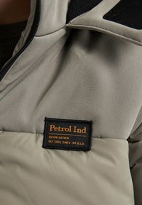 Giacca imbottita grigia con etichetta nera con "Petrol Ind" e testo arancione. Tessuto liscio con un mix di trame intorno al colletto.