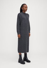 Πλεκτό μακρύ γκρι φόρεμα με ψηλό λαιμό και ριμπ μανσέτες, συνδυασμένο με μαύρες χοντρές γόβες μέχρι τον αστράγαλο. Απαλή υφή, ευθεία σιλουέτα.