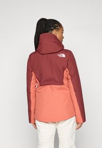 Tofarget vanntett jakke med maroon øverdel og korall underdel, med hette og merket logo på baksiden. Glatt tekstur.