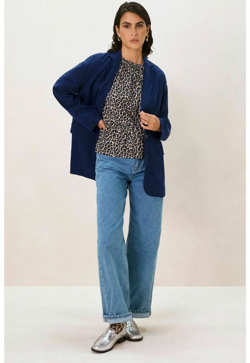 Blazer marine sur un chemisier à imprimé léopard, associé à un jean large bleu clair. Mocassins argentés, bracelets visibles et une bague. Fond neutre.