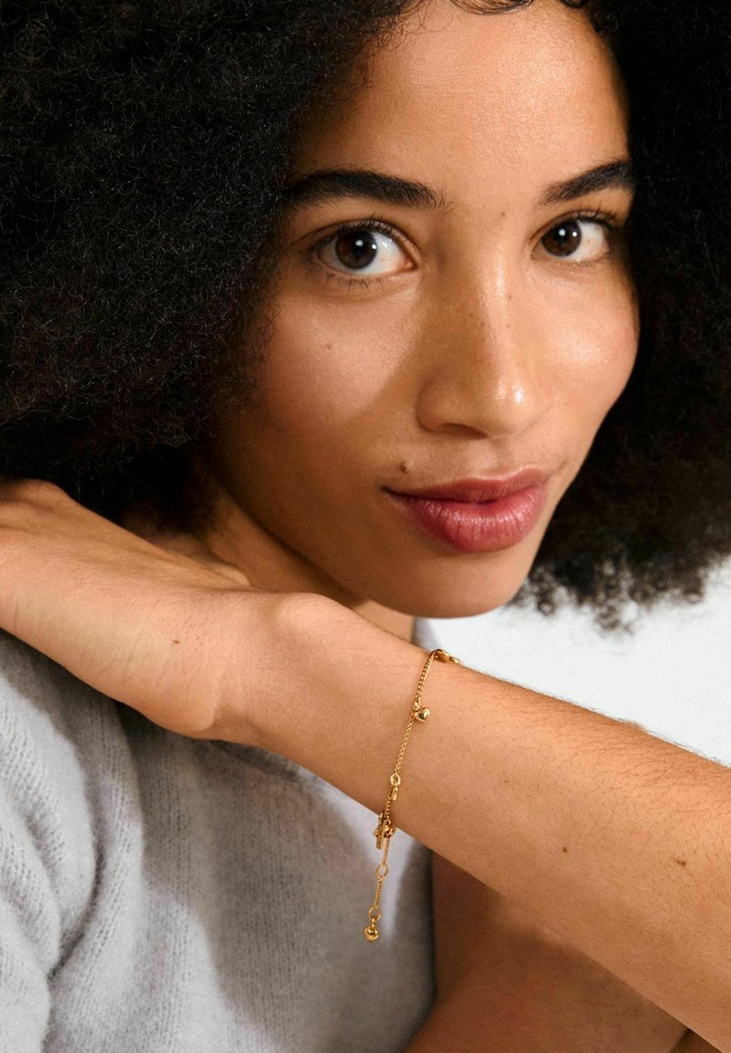Guldbaserad armband med runda och dekorativa hängande detaljer, bärs på handleden. Designen har en delikat kedja med varierande länkstorlekar och former.