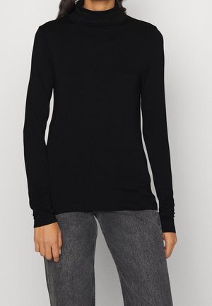 Kvinde iført en tætsiddende sort turtleneck med lange ærmer og falmede mørke grå jeans, stående mod en ensfarvet baggrund.