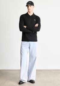 Črna polo majica z dolgimi rokavi, ovratnikom in zadrgo do polovice, kombinirana s širokimi svetlo modrimi kavbojkami in črnimi čevlji brez zavezovanja. Črna kapa z belim logotipom.