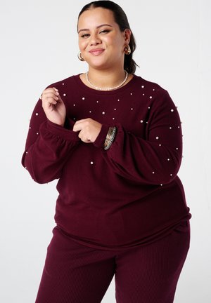 Vrouw in maroon trui met parelversieringen en bijpassende geribbelde broek past haar mouw aan, terwijl ze gouden hoepeloorbellen, armbanden en een parelketting draagt.