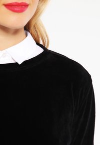 Donna bionda con rossetto rosso che indossa una camicia bianca con colletto sotto un top di velluto nero, su uno sfondo bianco.