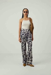 Mulher vestindo uma blusa branca sem mangas, calças azul-escuro com padrão floral branco, sapatos marrons de bico fino, óculos de sol e bolsa de ombro marrom.