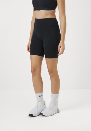Femme portant un short de sport noir taille haute, un soutien-gorge de sport noir, des chaussettes blanches Nike et des chaussures de sport blanches, debout sur un fond uni.