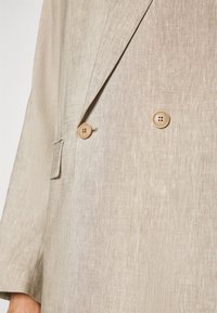 Beige linen blazer with a double-breasted design, featuring two round beige buttons and a textured fabric. Pockets visible on the sides.