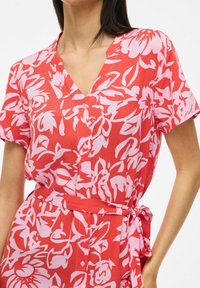 Abito rosso con stampa floreale, maniche corte, scollatura a V, chiusura con bottoni e vita con cintura annodata. Realizzato in tessuto leggero.