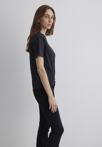 Black short-sleeve t-shirt with a rounded neckline, paired with fitted black jeans, displayed on a model standing sideways against a light background.