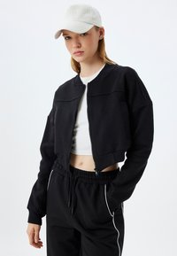 Sudadera negra recortada con cremallera frontal, puños acanalados y un cuello redondo, combinada con pantalones de chándal negros con ribetes blancos en los laterales.