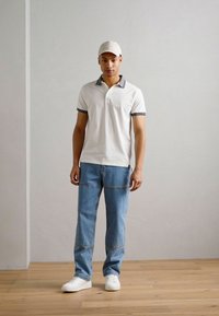 Polo alb cu detalii gri la guler, blugi largi de culoare albastru deschis și adidași albi; modelul stă în fața unui fundal simplu.