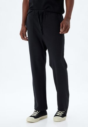 Sorte teksturerede bukser med elastisk talje og justerbar snøre, der har sidelommer, iført sorte sneakers med hvide såler.