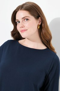 Marinblå långärmad skjorta med bred halsringning. Slät textur. Modellen bär guldiga hoops. Bakgrunden är helt vit.