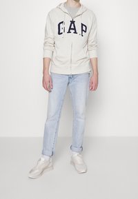 Felpa zipper beige chiaro con lettering navy "G.A.P.", t-shirt grigia sotto, jeans azzurri chiari e sneakers pallide con accenti grigi e bianchi.