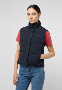 Gilet imbottito navy con colletto alto, bottoni a pressione e design trapuntato, dotato di tasche laterali e logo sul petto, abbinato a una t-shirt rossa.