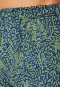 Tejido de colores azul y verde con un diseño de folia y aves, con una cinturilla elástica y un detalle de botón azul, etiquetado como "SCHIESSER."