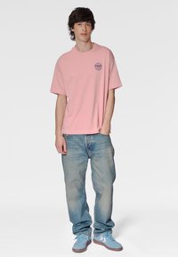 Ljusrosa bomullströja med rund halsringning och liten blå grafik på bröstet, matchad med bleka blåjeans och ljusblå sneakers.