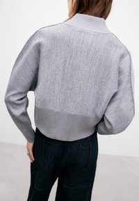 Grå kort sweater med en ribbet krave, tekstureret strik og lange ærmer. Båret over mørke, løstsiddende bukser.