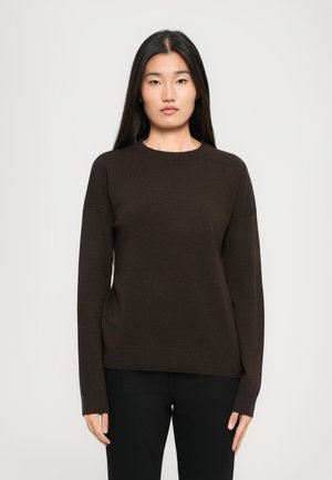 Femme avec de longs cheveux raides portant un pull marron foncé à manches longues et un pantalon noir, se tenant devant un fond clair et uni.