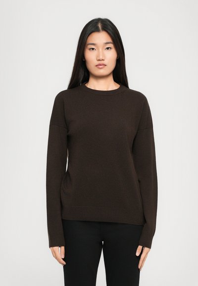 Femme avec de longs cheveux raides portant un pull marron foncé à manches longues et un pantalon noir, se tenant devant un fond clair et uni.