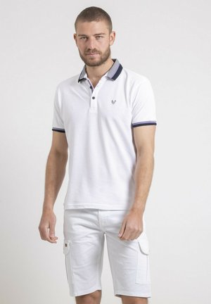 Homme portant un polo blanc à manches courtes avec un col foncé et un short cargo blanc, debout devant un fond clair uni.