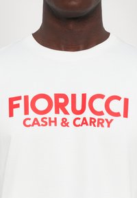 Tuvumā redzamā persona ar balto T-kreklu, uz kura krūtīm ir izteiksmīgs sarkans teksts "FIORUCCI CASH & CARRY".