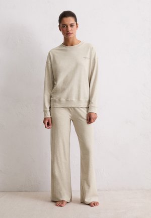 Donna in piedi a piedi nudi che indossa una felpa beige a maniche lunghe e pantaloni larghi abbinati contro un muro chiaro e semplice.