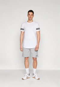 Valge T-särk mustade triipudega varrukatel, sobitatud hallide pükstega. Modell kannab valgeid tossusid beežide detailidega ja valgeid sokke.