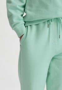 Mintgrüner Baumwollmischung-Trainingsanzug mit gerippten Bündchen, Kordelzugbund und Seitentaschen. Glatte Textur, entspannte Passform, minimale Designmerkmale.