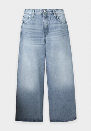 Mann mit dunkler Haut, der ein dunkelblaues kurzärmeliges Polohemd, hellblaue Jeans mit weitem Bein und schwarze Schuhe trägt, steht vor einem weißen Hintergrund.