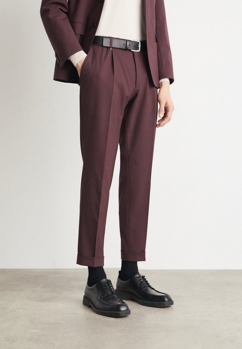 Burgundy tailored trousers with pleats, cuffed hem, paired with black leather shoes and a black belt, worn over a light sweater.