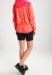 Veste légère et imperméable de couleur corail vif avec une capuche rose, associée à un short noir avec des détails perforés discrets. Baskets noires.