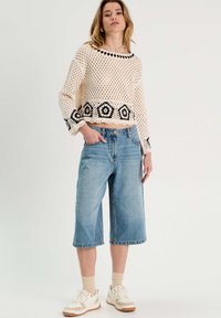 Suéter de crochet color crema con patrones florales negros, combinado con culotes de denim de pierna ancha color azul claro. La modelo lleva zapatillas ligeras y calcetines.