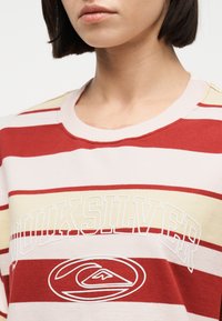 Γυναίκα που φοράει πουκάμισο με κόκκινες, λευκές και κίτρινες ρίγες και το κείμενο και το λογότυπο «Quiksilver» στο στήθος, ορατή από τον λαιμό έως το άνω μέρος του κορμού.