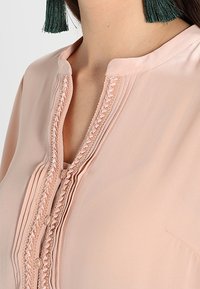 Primo piano di una persona che indossa una blusa di un rosa pallido con dettagli nei punti e bottoni, abbinata a orecchini con frange verdi.