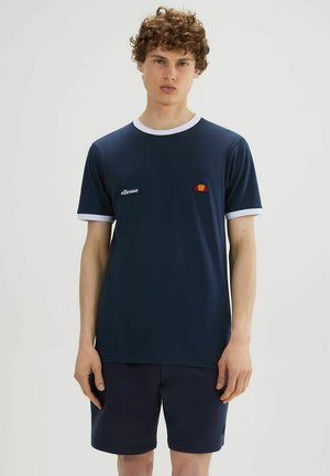 Młody mężczyzna z kręconymi włosami, ubrany w granatowy t-shirt z białymi lamówkami i logo, w połączeniu z granatowymi szortami, stojący na jednolitym tle.