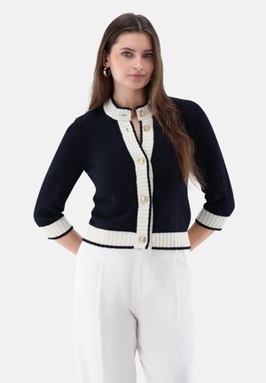 Woman with long hair wearing a black cardigan with white trim and gold buttons, paired with high-waisted white pants, posing with hands behind.