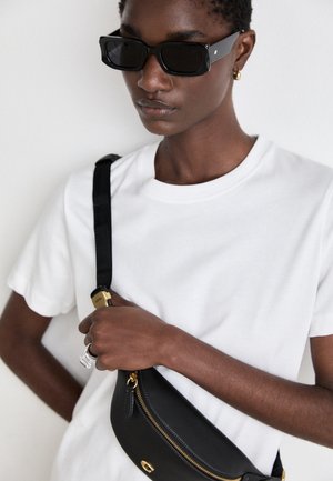Person iført sorte rektangulære solbriller, hvid T-shirt, guldring ørering, holder sort læder crossbody taske med gylden lynlås.