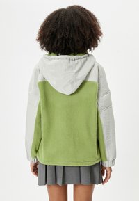 Chaqueta de forro polar verde con detalles en tela gris, capucha y puños elásticos, que presenta una textura suave y un corte relajado. Se muestra la vista trasera.