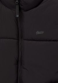 Veste matelassée noire avec une fermeture éclair à l'avant et un petit logo sur la poitrine gauche.